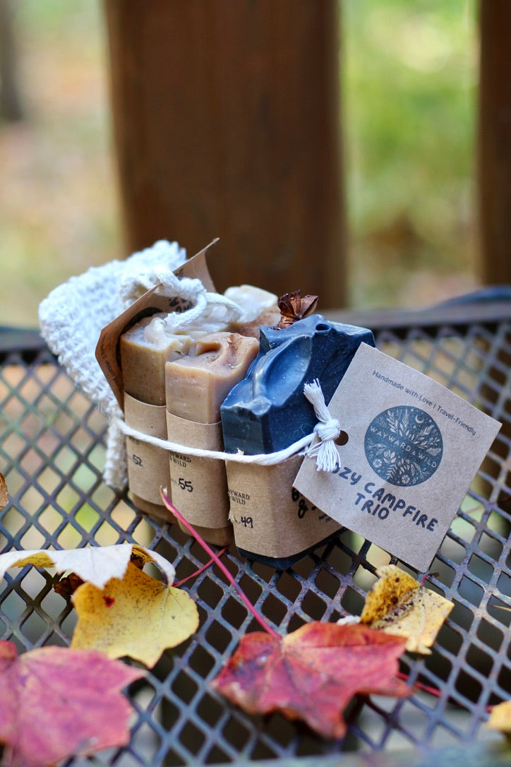 3 autumn-scented soaps sit on a grate surrounded by red and yellow leaves with a forested background.